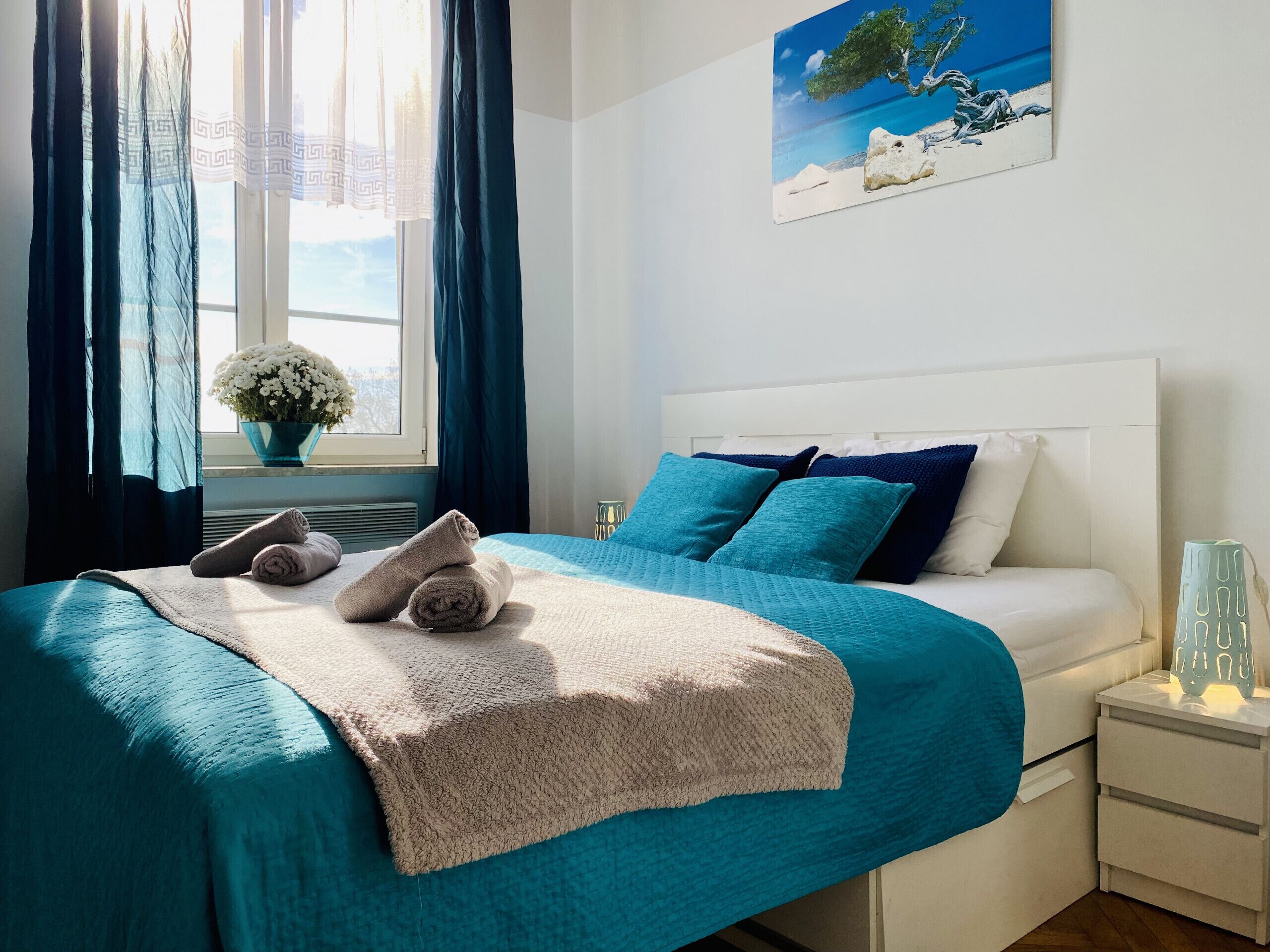 Un lit bien fait, recouvert d’une couvre-lit turquoise et d’un plaid beige, une table de chevet blanche avec une lampe, et un tableau représentant une scène de plage accroché au mur.