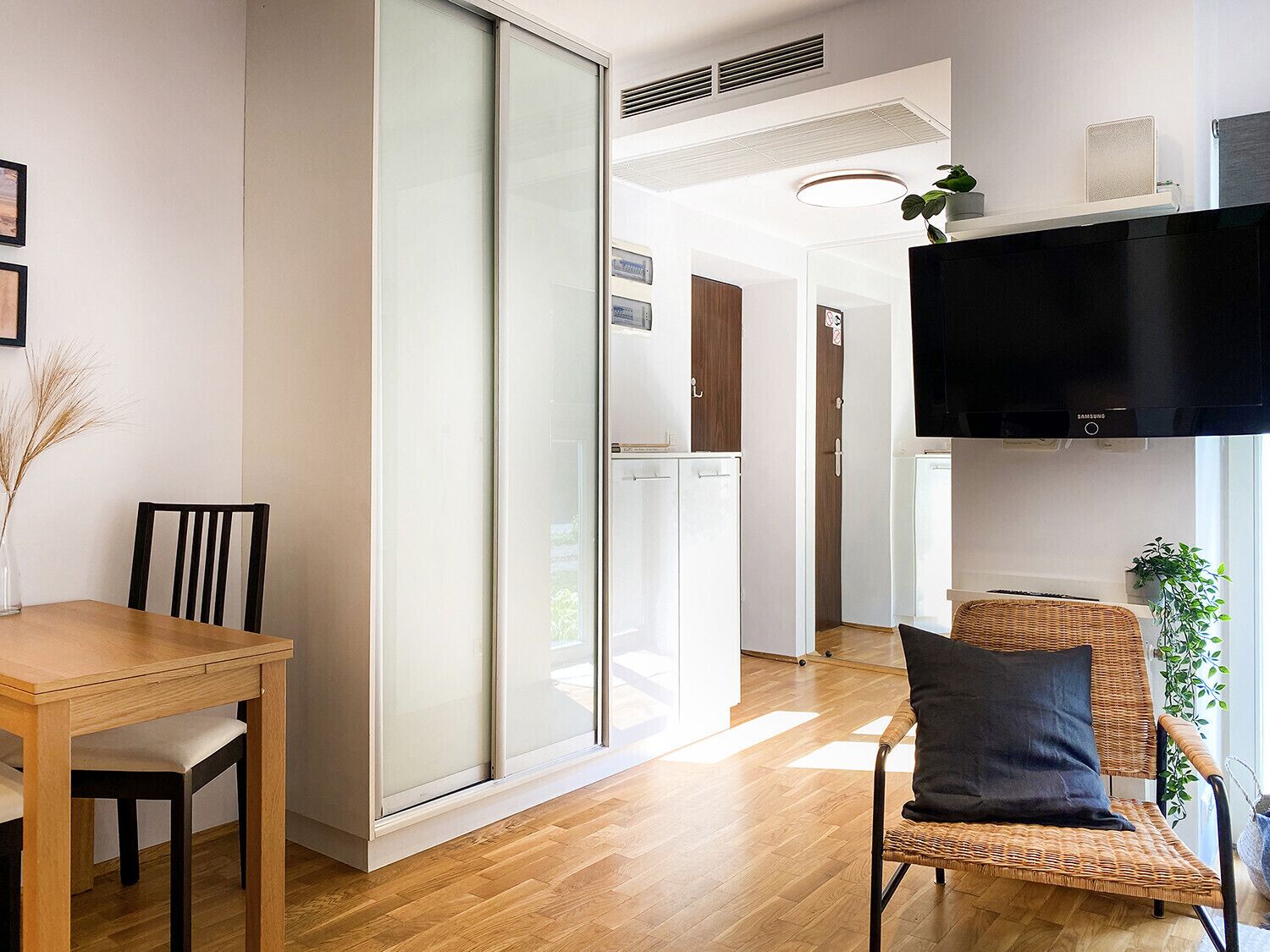 A modern living room with a wooden dining table, a wicker chair with a cushion, a large flat-screen TV mounted on the wall, and a sliding glass door leading to another room.