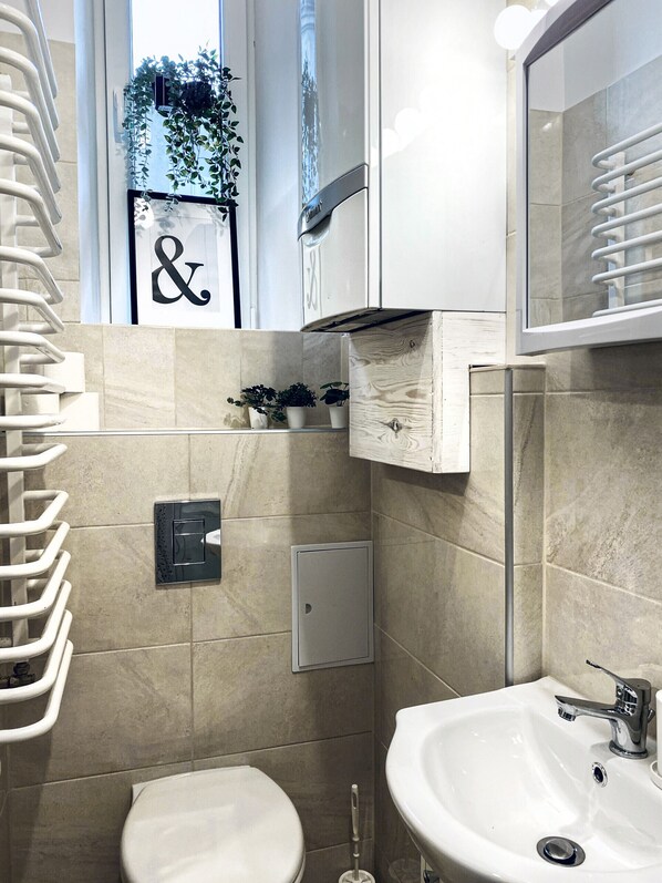 Et moderne badeværelse med toilet, vask og håndklæder. Der er et lille skab med planter og et skilt med et symbol.