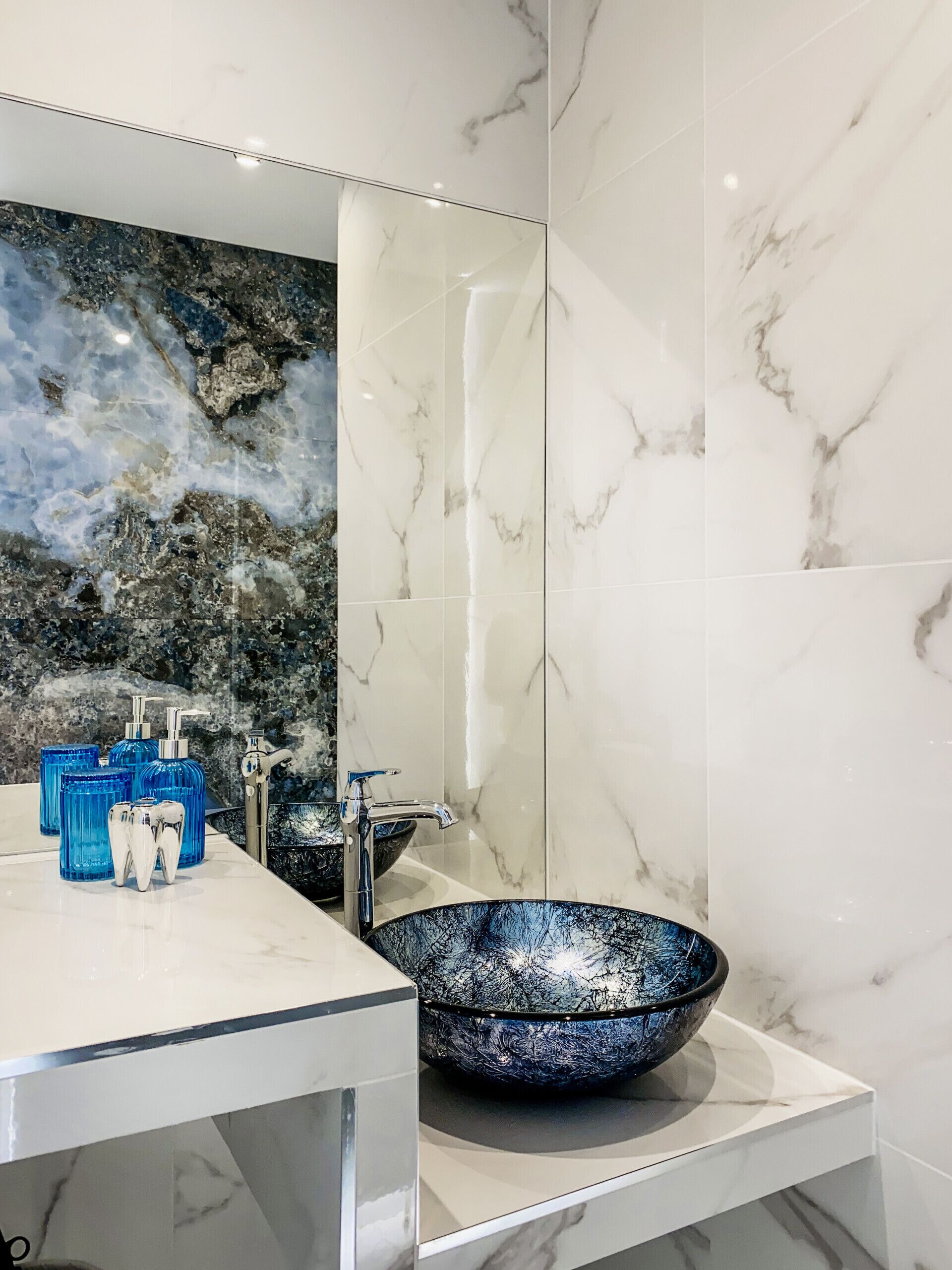 Baño con tocador de mármol, un espejo grande, lavamanos de vidrio azul y botellas de artículos de tocador azules.