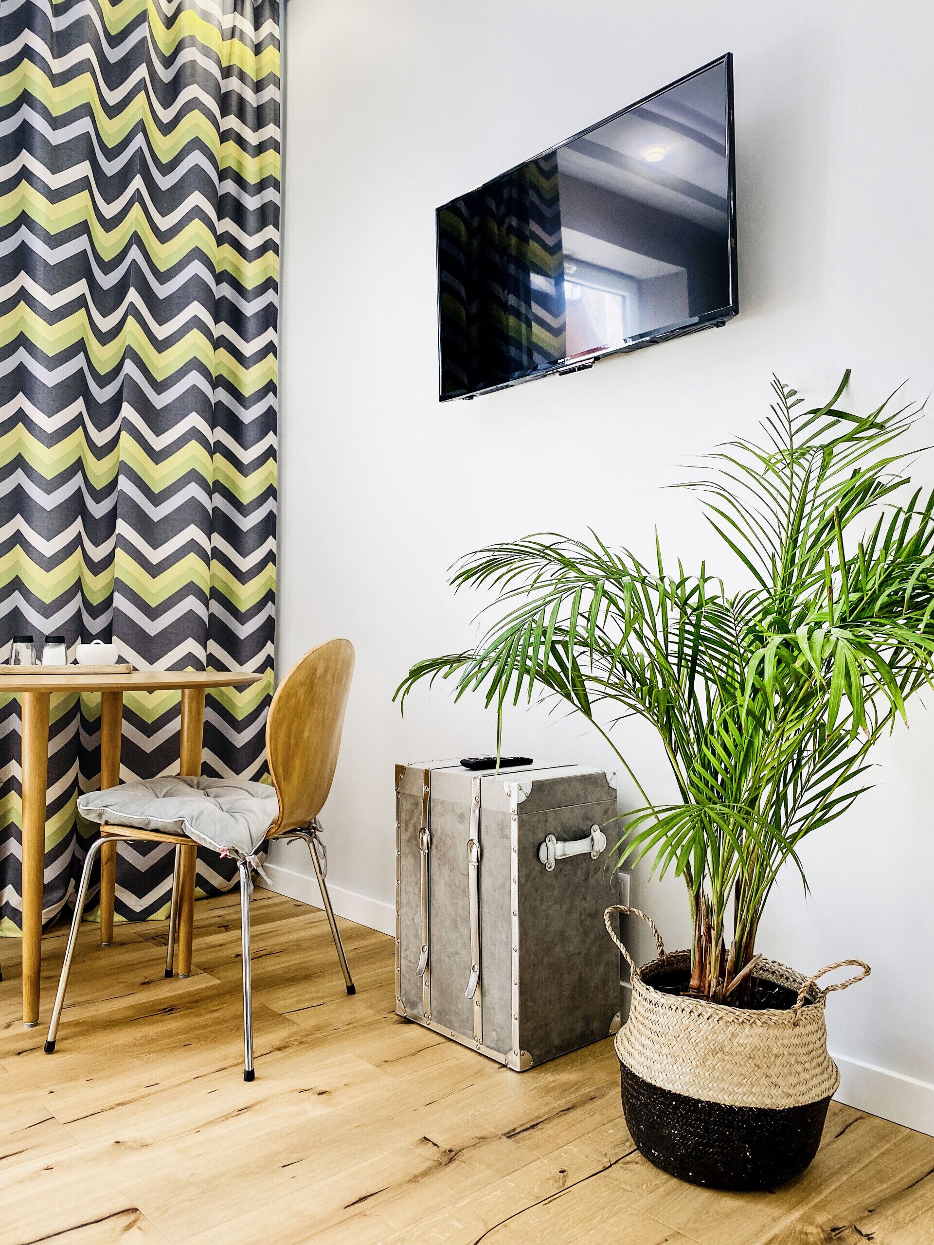 Een kamer met houten vloer, een aan de muur bevestigde televisie, een kamerkleurig gordijn, een houten tafel met een stoel, een koffer en een bloempot.