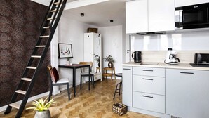 A modern kitchen with white cabinets, a black microwave, and a coffee maker. There is a wooden dining table with chairs, a ladder against the wall, and a patterned wallpaper on one side.