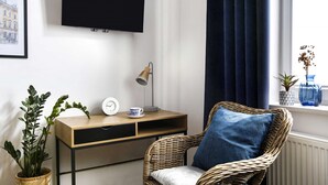 Habitació amb televisió penjada, taula auxiliar de fusta amb un rellotge i una tassa, cadira de vimetlla amb coixí, una planta en test i una finestra amb cortines.