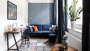 A modern living room with a blue sofa, a small table with a lamp, and a potted plant by the window.