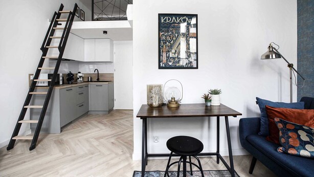 A modern kitchen with a wooden ladder, a dining area with a table and chair, and a sofa with cushions.