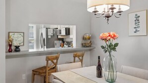 A dining area with a table, chairs, a vase with flowers, a chandelier, and a kitchen in the background.