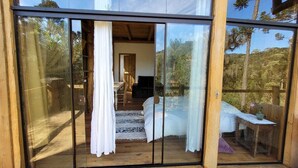 A wooden cabin with a large sliding glass door leading to a bedroom with a bed, a chair, and a small table. - YAAHOO ECOLODGE (Bocaina do Sul)