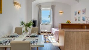 A room with a dining table set for two, a small seating area with a sofa, a TV, and a view of the sea through a window. - Apartment & Studio Riva Lucija by Litto (Split)