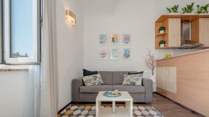 A modern living room with a grey sofa, a coffee table, and a patterned rug. There are framed pictures on the wall, a window, and a kitchenette area with cabinets and plants. - Apartment & Studio Riva Lucija by Litto (Split)