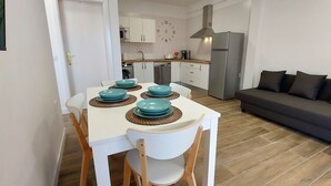 Un espace de vie compact comprenant un coin repas avec une table blanche et quatre assiettes bleues, un canapé gris et une cuisine équipée d’appareils électroménagers modernes.
