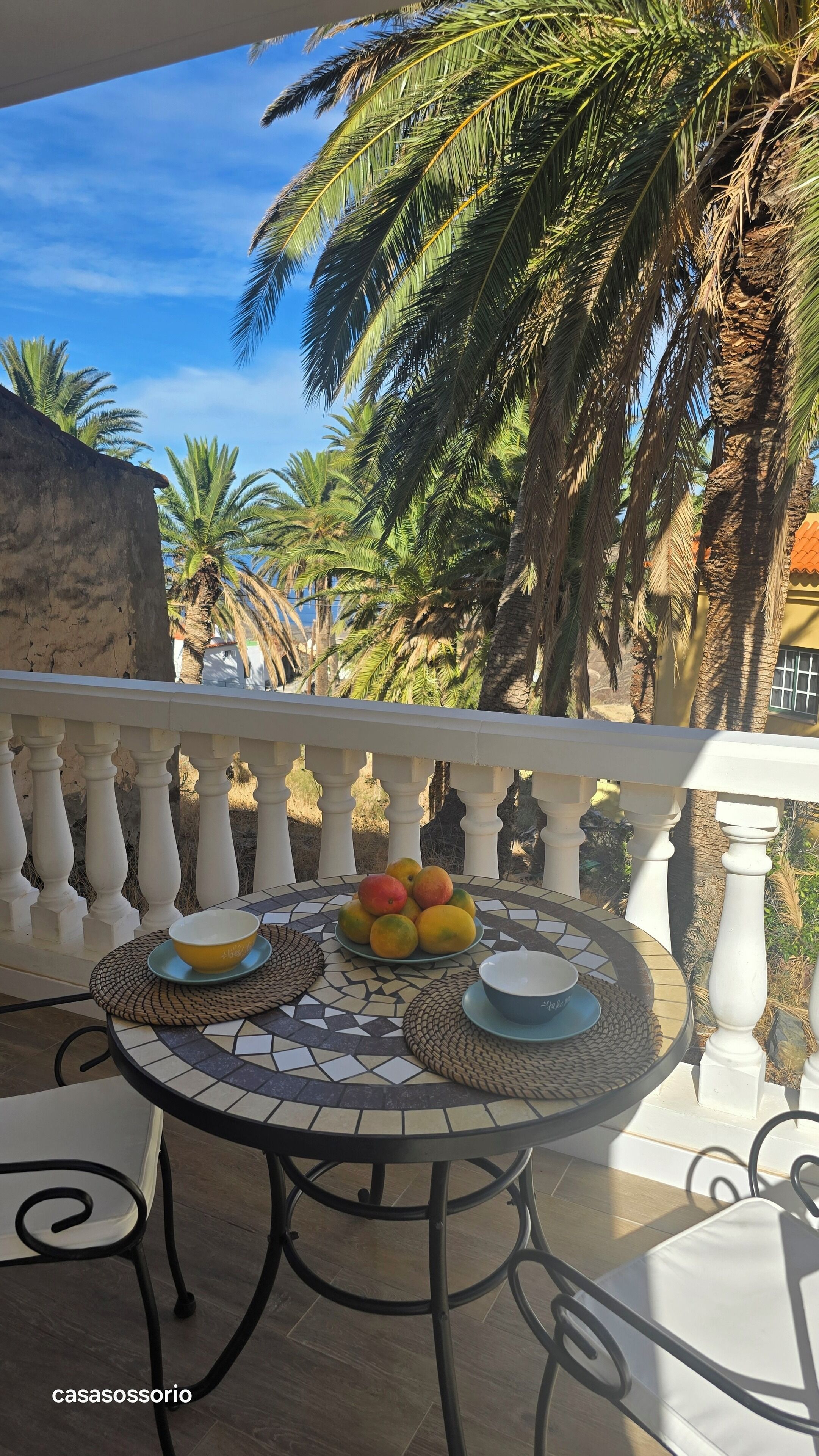 A round table with a mosaic top, two bowls, and a plate of fruit on a balcony with a white balustrade and palm trees in the background.