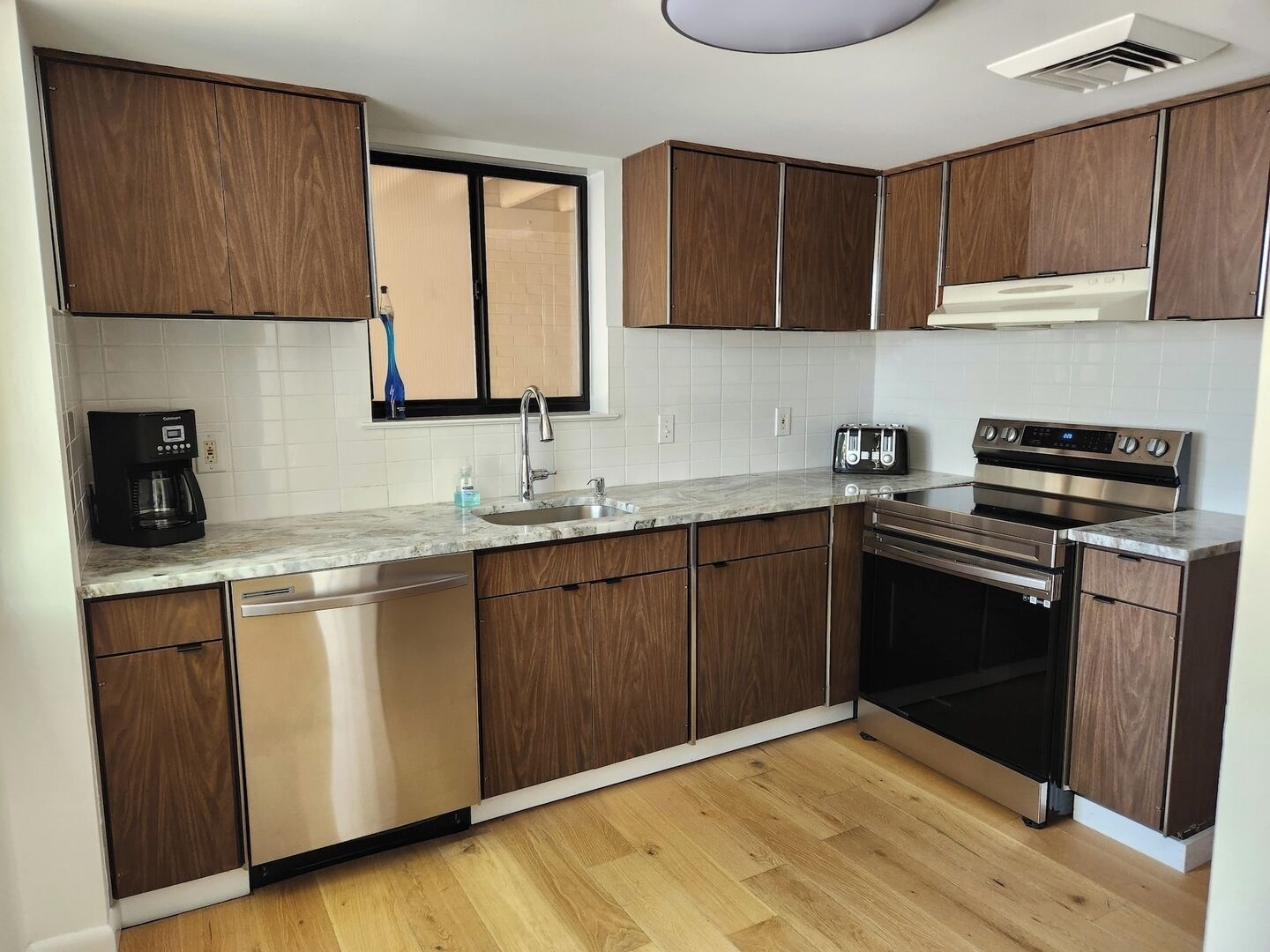 A modern kitchen with wooden cabinets, a built-in dishwasher, a coffee maker, a toaster, and a stainless steel oven.