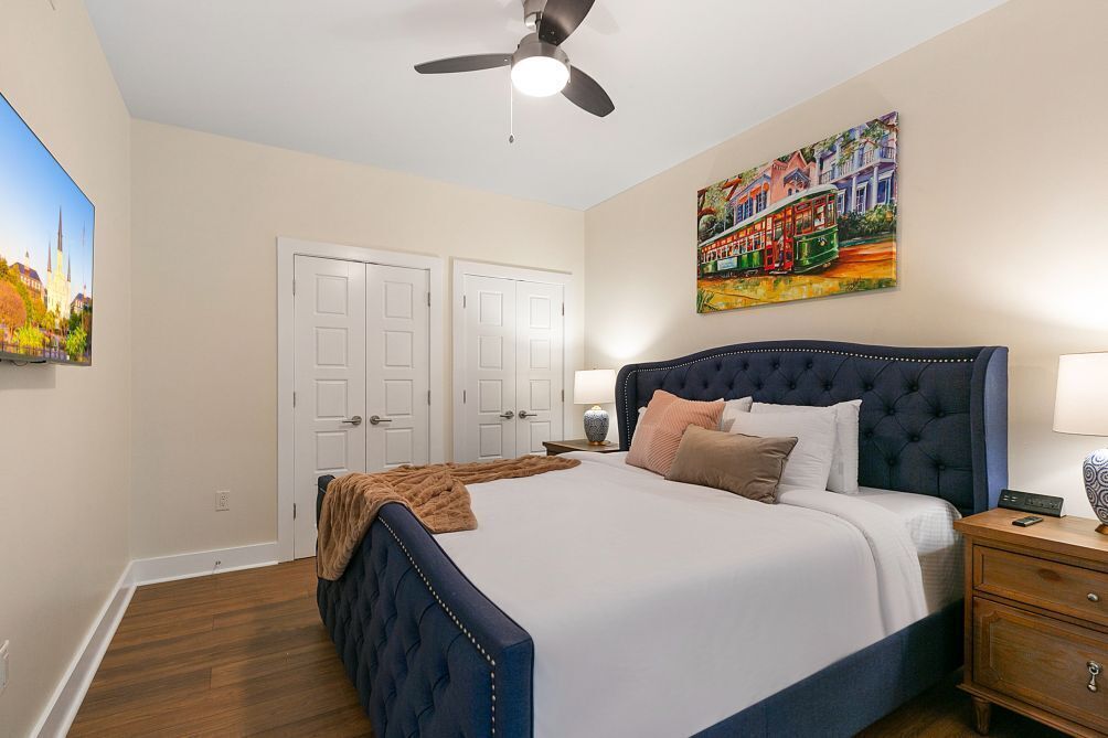A bedroom with a large bed, a ceiling fan, two white doors, a painting of a tram, and a nightstand with a lamp.