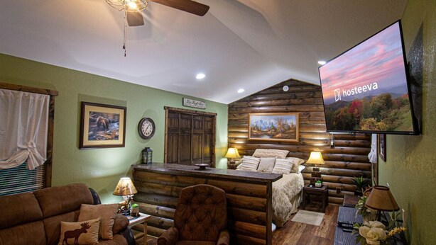 A cozy log cabin-style room with a bed, a chair, a sofa, and a television displaying 'Hosteeva'. - The Lil House with Balcony (Robbinsville)