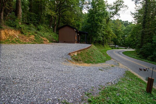 A gravel parking area with a wooden building, a curved road, and dense greenery. - The Lil House with Balcony (Robbinsville)