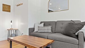 A modern living room with a grey sofa, a wooden coffee table, a small side table, a wall-mounted mirror, and a sign that reads 'PRAY FOR SURF'. - Manresa Surf House 1 bed upstairs unit #4 (La Selva Beach)