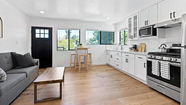 A modern kitchen with white cabinets, a stainless steel stove, and a dining area with a wooden table and chairs. - Manresa Surf House 1 bed upstairs unit #4 (La Selva Beach)