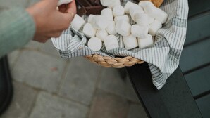 A person holding a stick with marshmallows and chocolate pieces, with a basket of marshmallows and chocolate pieces in the foreground.