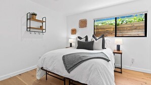 A bedroom with a bed, two bedside tables, a window with a view, a wall-mounted shelf, and a ceiling light. - Manresa Surf House 1 bedroom Sleeps 4 unit #2 (La Selva Beach)