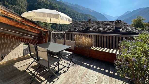 Eine Terrasse mit Tisch und Stühlen, einem weißen Sonnenschirm und Blick auf Berge.