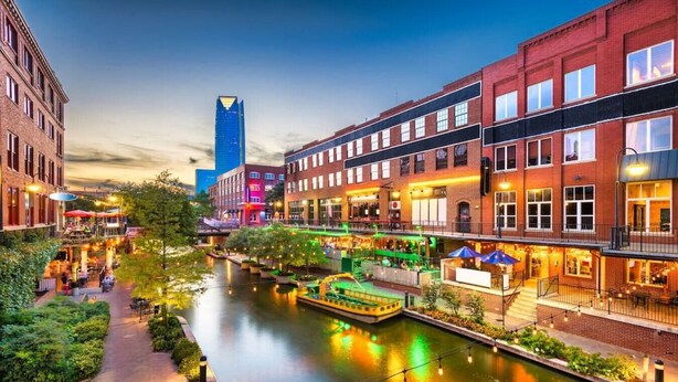 A riverside walkway with buildings, a boat, and outdoor seating areas. - "The Cozy Cabana" in Paseo (Oklahoma City)