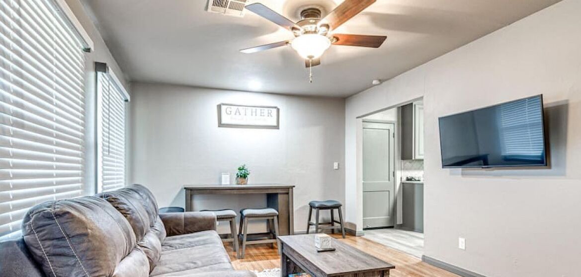 A living room with a sofa, coffee table, and a ceiling fan. There is a sign on the wall that says "GATHER".
