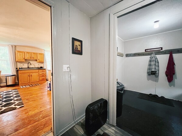 A room with a wooden floor, a black fan, a white door, and a kitchen visible through an open door. - The Olde Farmhouse - Historic Property - 10 miles from Cherry Springs State Park (Coudersport)