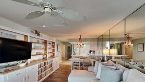 A spacious living room with a large flat-screen TV, a white sofa, and a dining area with a chandelier. - Regency Towers by Resort Collection (Panama City Beach)