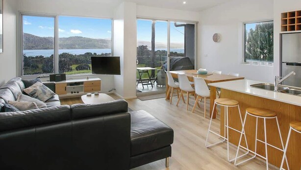 A modern living space with a sofa, dining area, kitchen, and large windows offering a view of the outdoors. - Bay Vue - Absolute Oceanviews! (Apollo Bay)