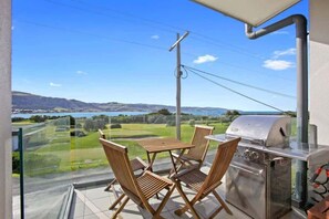A balcony with a grill, chairs, and a view of a lake and hills.