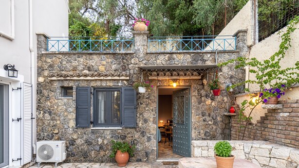 Un edificio de piedra con una puerta, plantas en macetas y un pequeño espacio al aire libre con mesas y sillas.