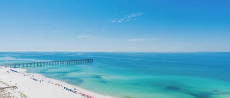 A beach with a pier extending into the water, clear turquoise water, and a row of beach umbrellas.