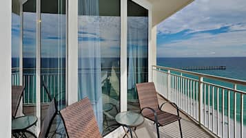 A balcony with wicker chairs and a glass table, overlooking a beach and a pier.