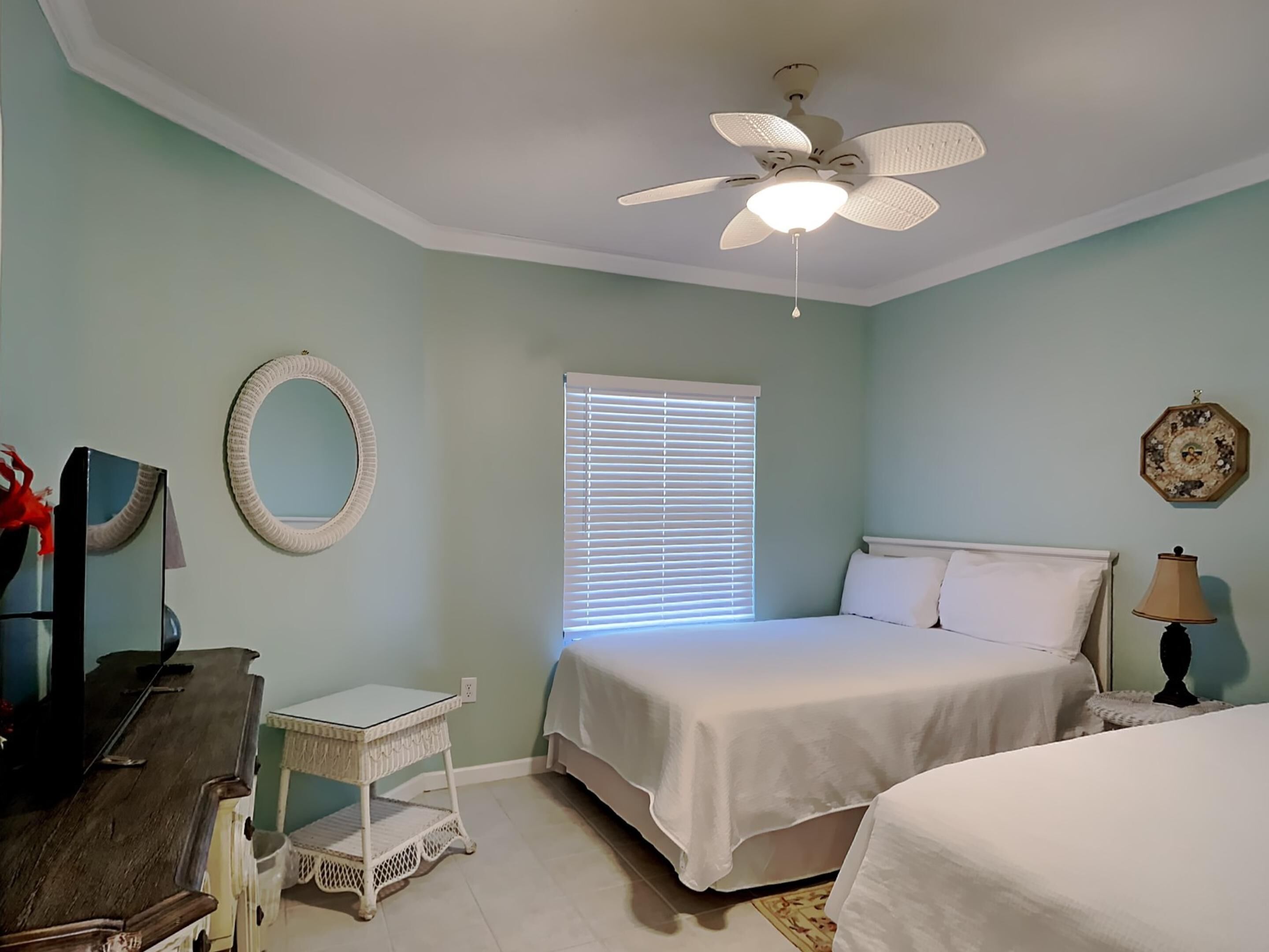 A bedroom with a bed, a ceiling fan, a window with blinds, a small table, and a lamp.