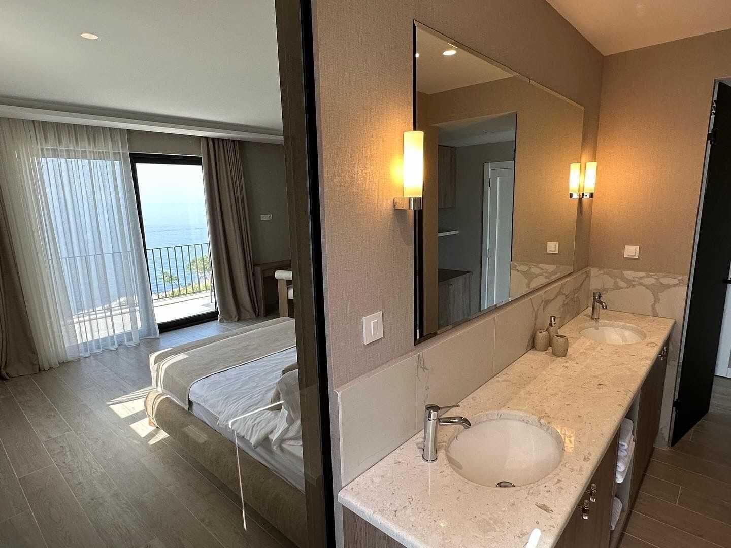 Una habitació d'hotel amb un llit gran, un bany amb un lavabo i mirall, i un balcó amb cortines.