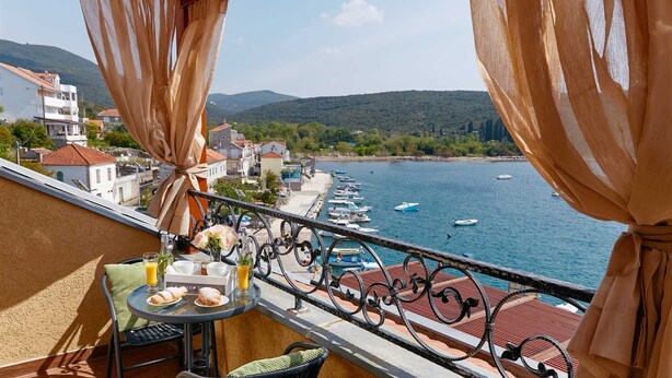 Ein Balkon mit einem Frühstückstisch, der auf einen Yachthafen mit Booten und eine hügelige Landschaft im Hintergrund blickt.