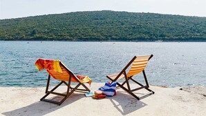 Zwei Strandstühle mit Handtüchern an einem sandigen Ufer, ausgerichtet auf einen stillen See mit einem bewaldeten Hügel im Hintergrund.