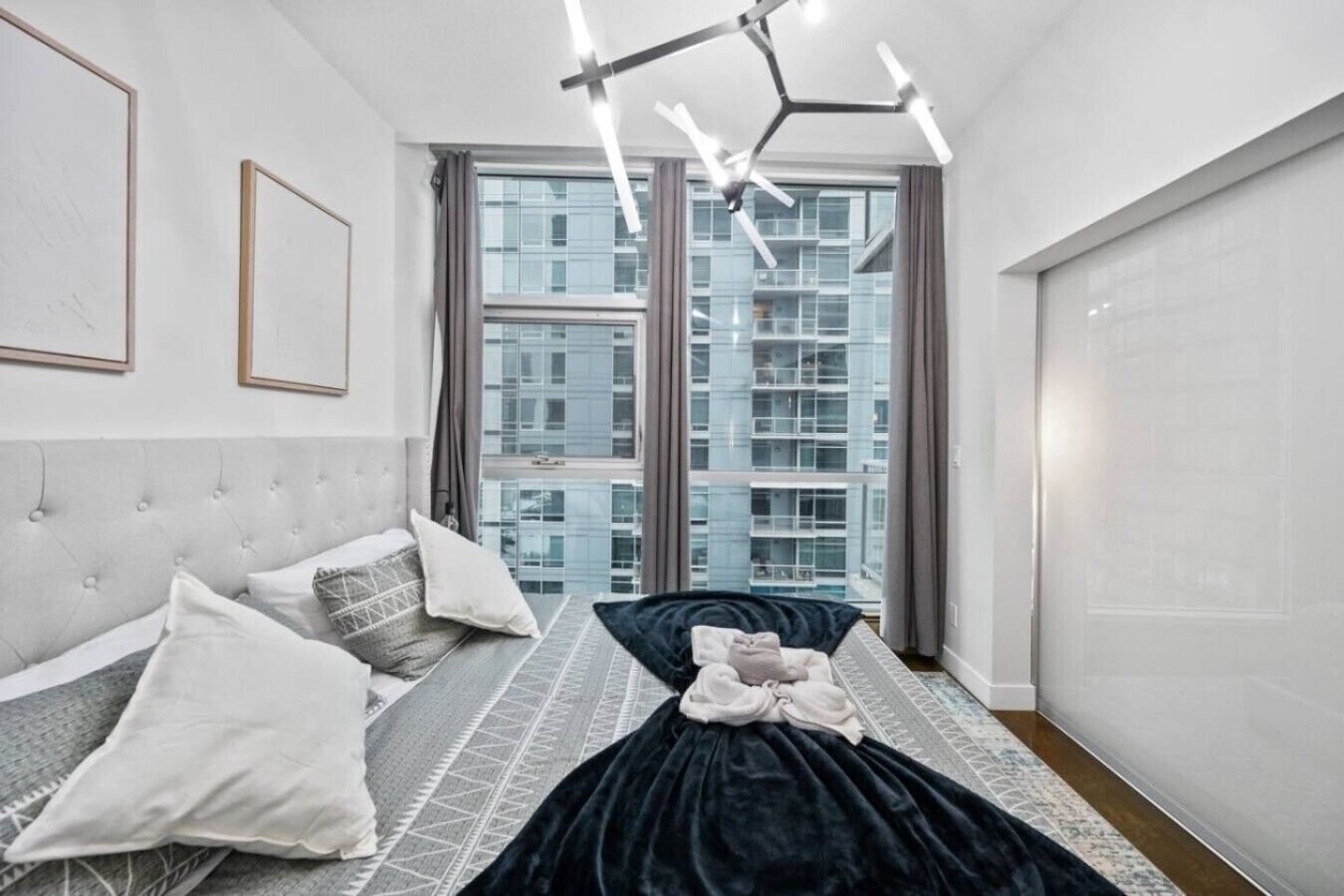 A modern bedroom with a tufted headboard, a bed with a dark cover, a patterned rug, and a view of high-rise buildings through large windows.