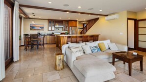 A spacious living area with a large sofa, a coffee table, and a dining area with chairs and a kitchen in the background.