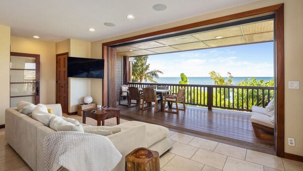 A spacious living room with a large sofa, a coffee table, and a TV. There is a balcony with a view of the ocean.