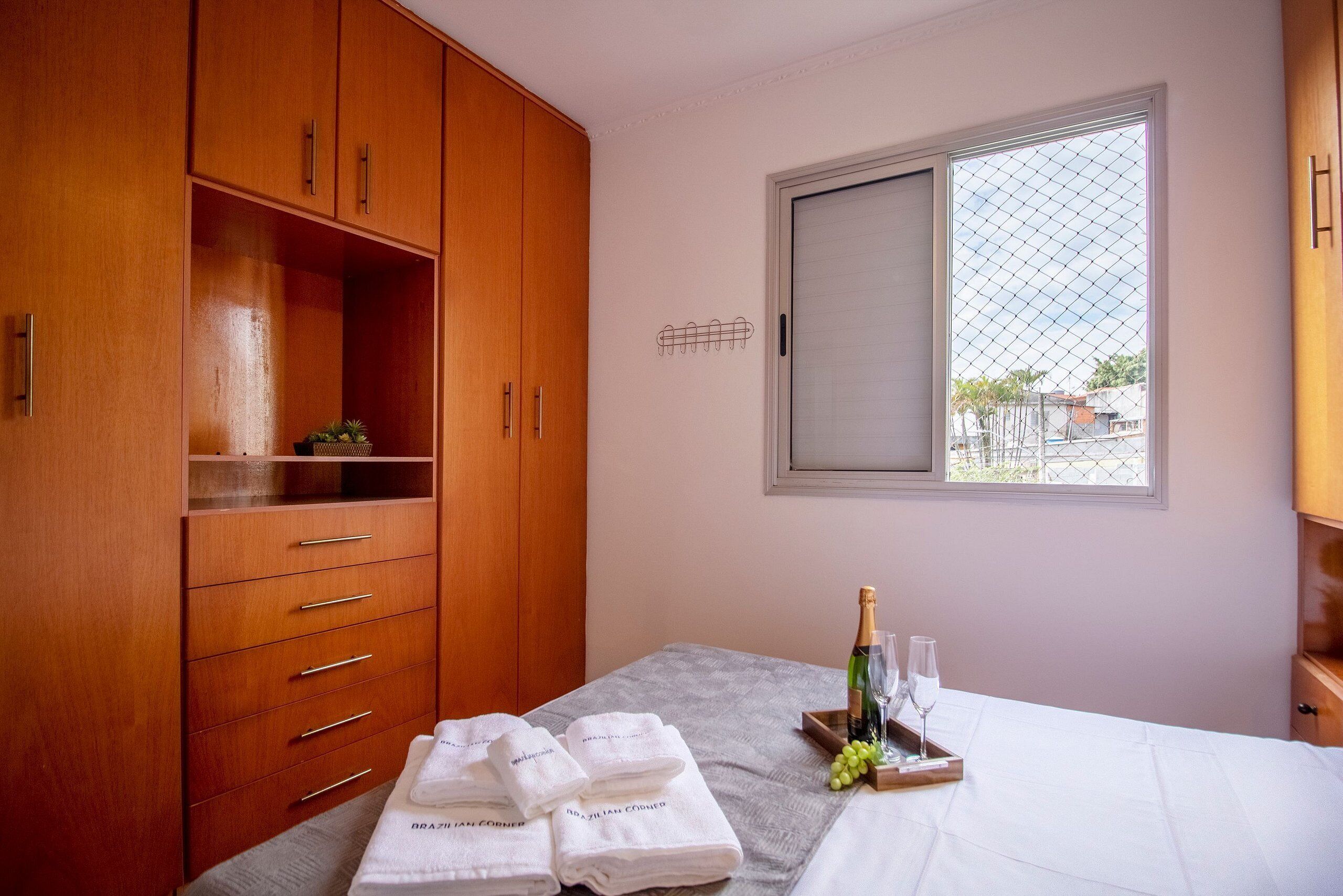 Een hotelkamer met een houten garderobe, een bed met witte lakens, een dienblad met een fles en glazen, en een raam met uitzicht op gebouwen.