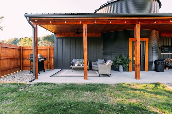 Un patio cubierto con mobiliario exterior, una parrilla y un depósito cilíndrico grande al fondo.