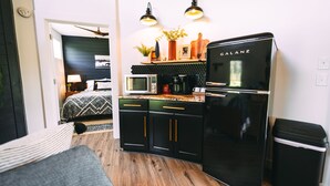 A compact kitchen with a black refrigerator, microwave, and coffee maker. There are two pendant lights above the counter, and a small trash bin to the right.