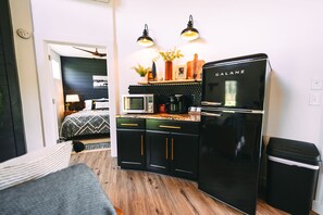 A compact kitchen with a black refrigerator, microwave, and coffee maker. There are two pendant lights above the counter, and a small trash bin to the right.