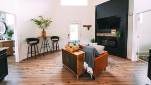 A modern living room with a wooden floor, a black wall unit, a sofa, and a dining area with stools and a table.
