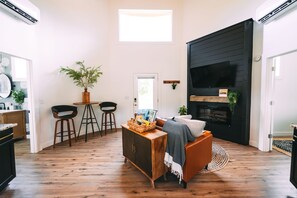 A modern living room with a wooden floor, a black wall unit, a sofa, and a dining area with stools and a table.