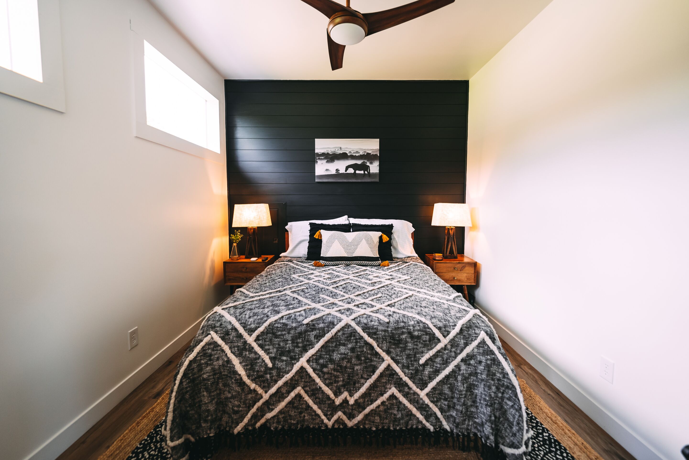 A bedroom with a bed centered under a ceiling fan, flanked by two bedside tables with lamps, a dark headboard, and a framed picture above the bed.