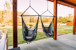 Two gray hammocks suspended from a wooden structure with a view of a grassy yard and trees.