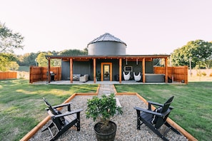 Un patio cubierto con mobiliario exterior y un gran silo de grano al fondo.