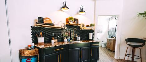 Una cocina moderna con armarios negros, una encimera de madera y un salpicadero negro. Hay dos luces empotradas, una cesta de mimbre y una silla.